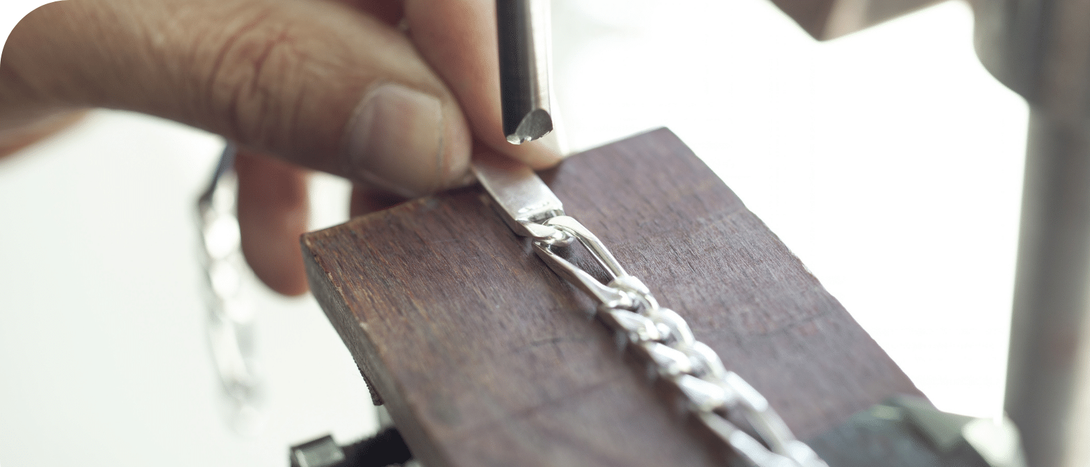 Main d'artisan travaillant une chaîne en argent sur un établi en bois, à l'aide d'un outil
